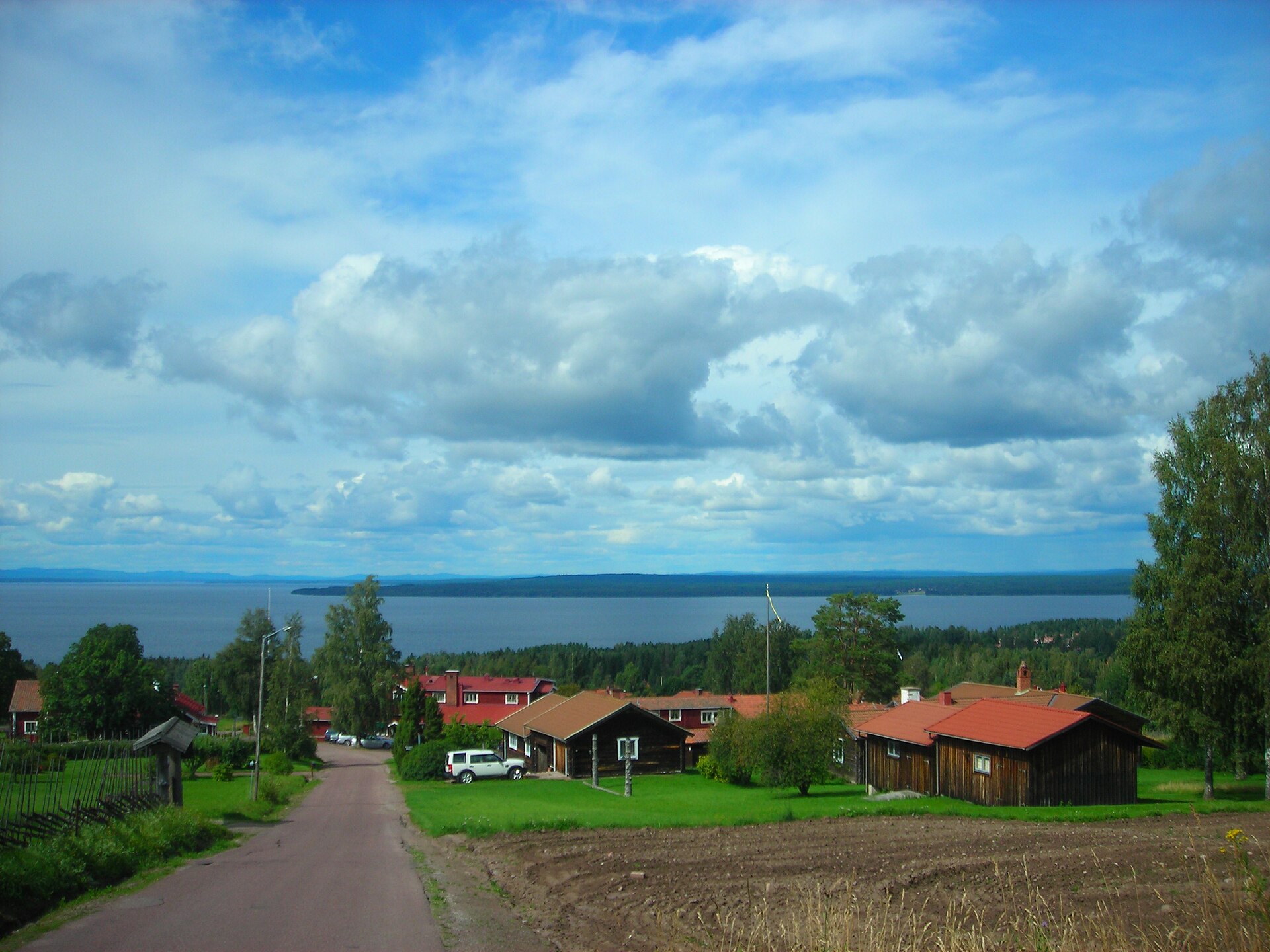 Dalarna: Holzhäuser in Tällberg am Hang, dahinter der Siljansee unter Sommerwolken