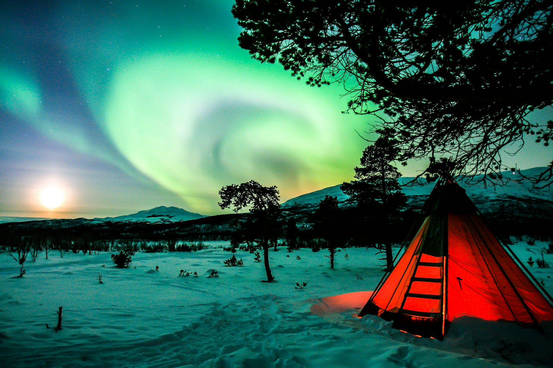 Lappland Urlaub im Winter: Polarlicht über verschneiter Landschaft bei Abisko mit beleuchtetem Zelt