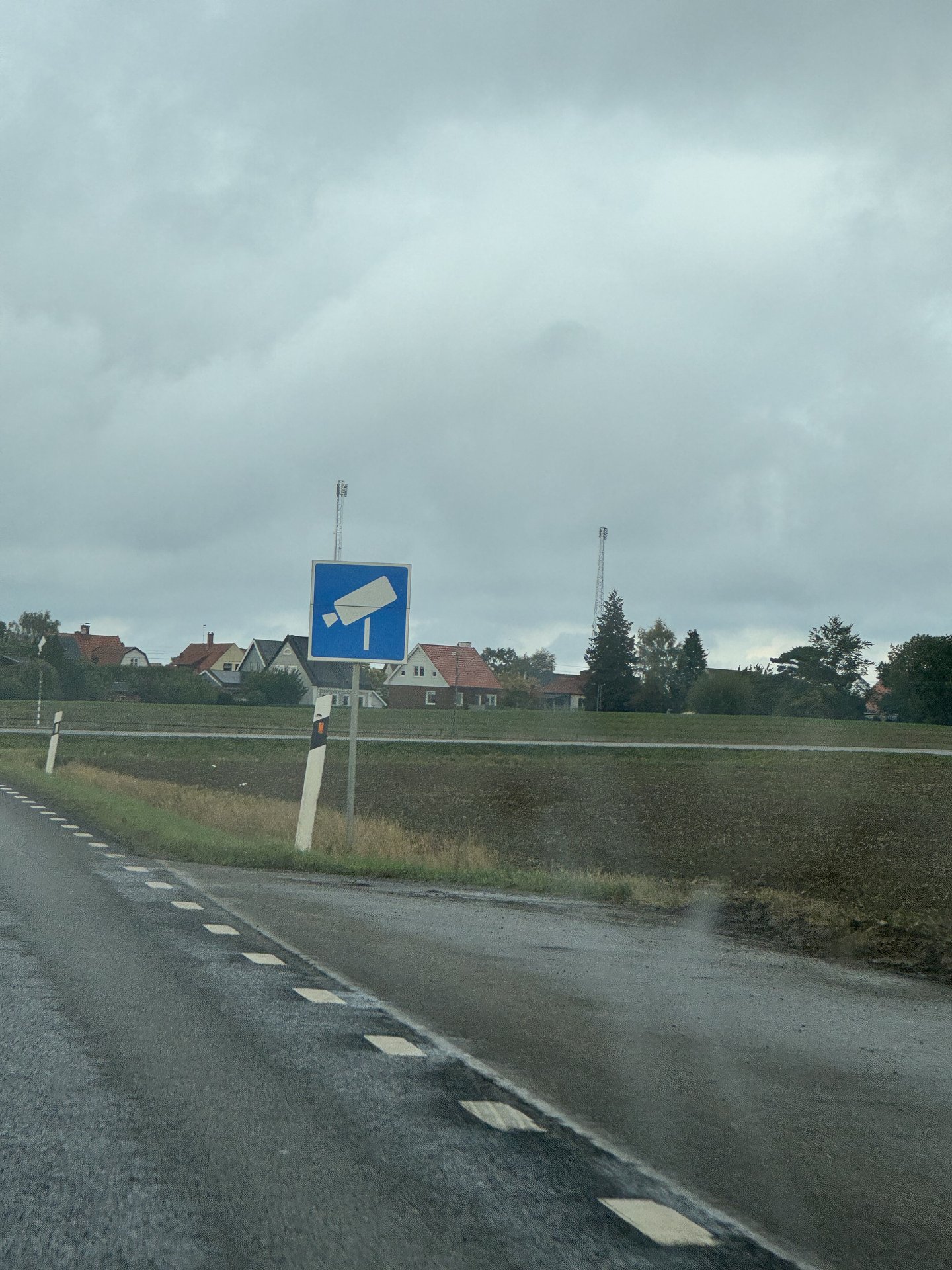 Blaues Hinweisschild mit weißem Kamerasymbol am Rand einer Landstraße in Skåne, dahinter Felder und Häuser