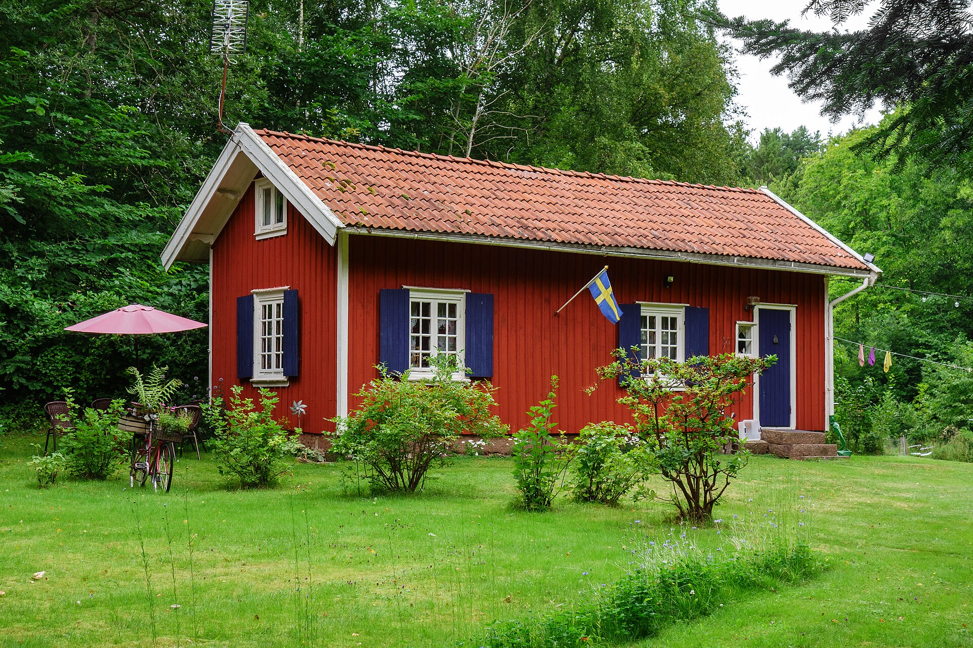 Hütte in Schweden: rote Holzhütte mit weißen Ecken und blauen Fensterläden auf einer Wiese am Waldrand bei Brastad in Bohuslän