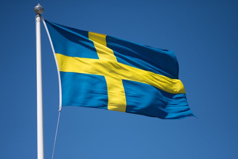 Schwedische Flagge in Blau und Gelb weht im Wind an einem weißen Fahnenmast vor blauem Himmel