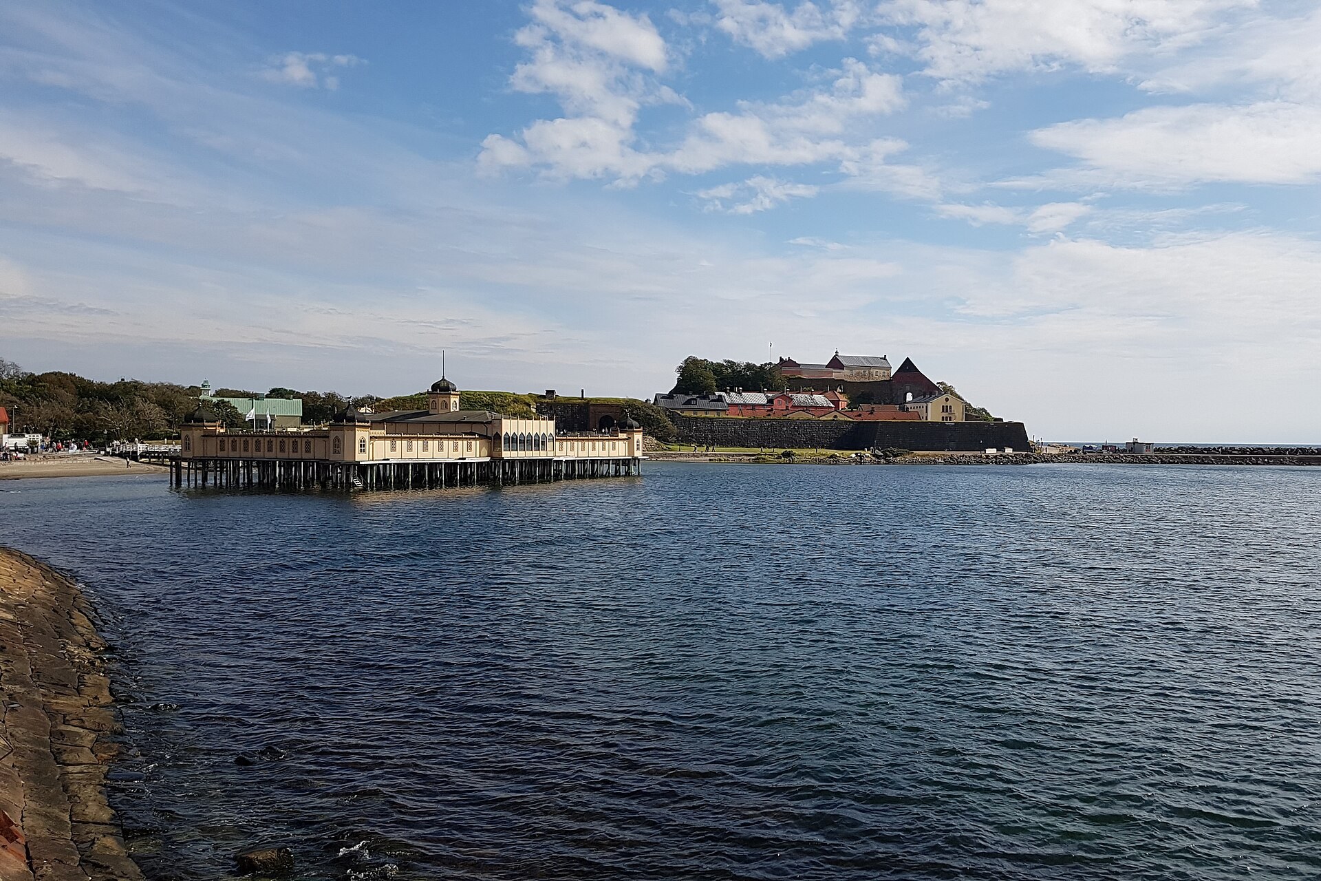 Das Kaltbadehaus auf Pfählen im Meer und die Festung Varberg im Hintergrund unter blauem Himmel